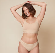 Carli is standing facing forward with her arms raised above her head, smiling. She is wearing the Bliss Racerback Bralette and the Bliss Bikini in Beige, showcasing the set from the front.