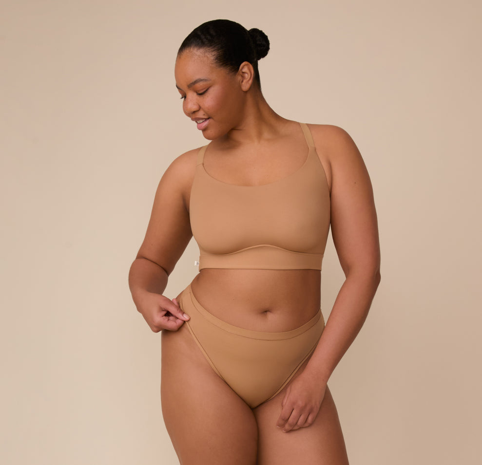 Zarria stands casually with a soft smile wearing the Bliss Racerback Bralette and Bliss Highwaist Thong in Tan. The scoop neckline and wide band of the bralette pair with the high-rise thong’s soft microfiber fabric, offering comfort and a seamless look.