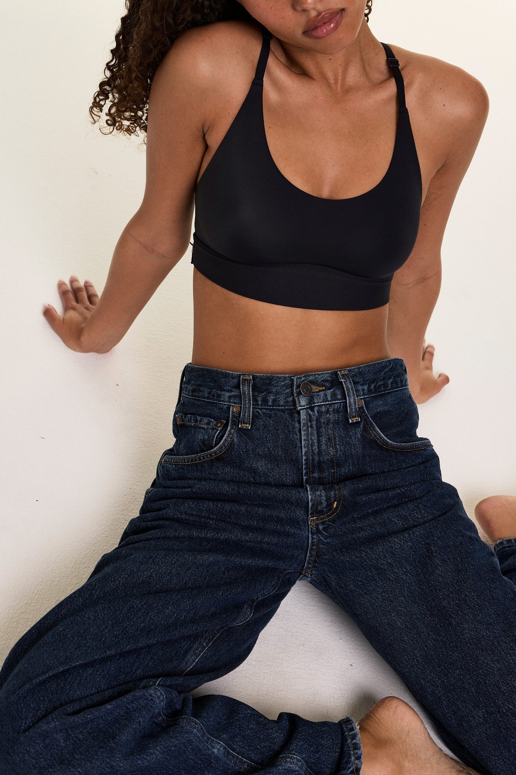 In a studio setting, Courtney wears the Bliss Racerback Bralette in Black with relaxed-fit jeans, leaning against a wall to showcase the scoop neckline and smooth fabric.