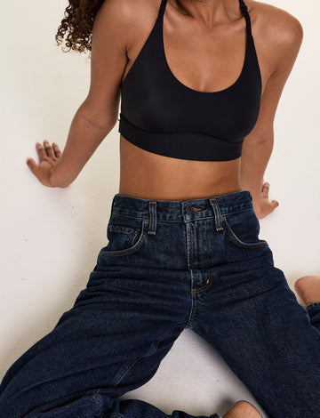 In a studio setting, Courtney wears the Bliss Racerback Bralette in Black with relaxed-fit jeans, leaning against a wall to showcase the scoop neckline and smooth fabric.