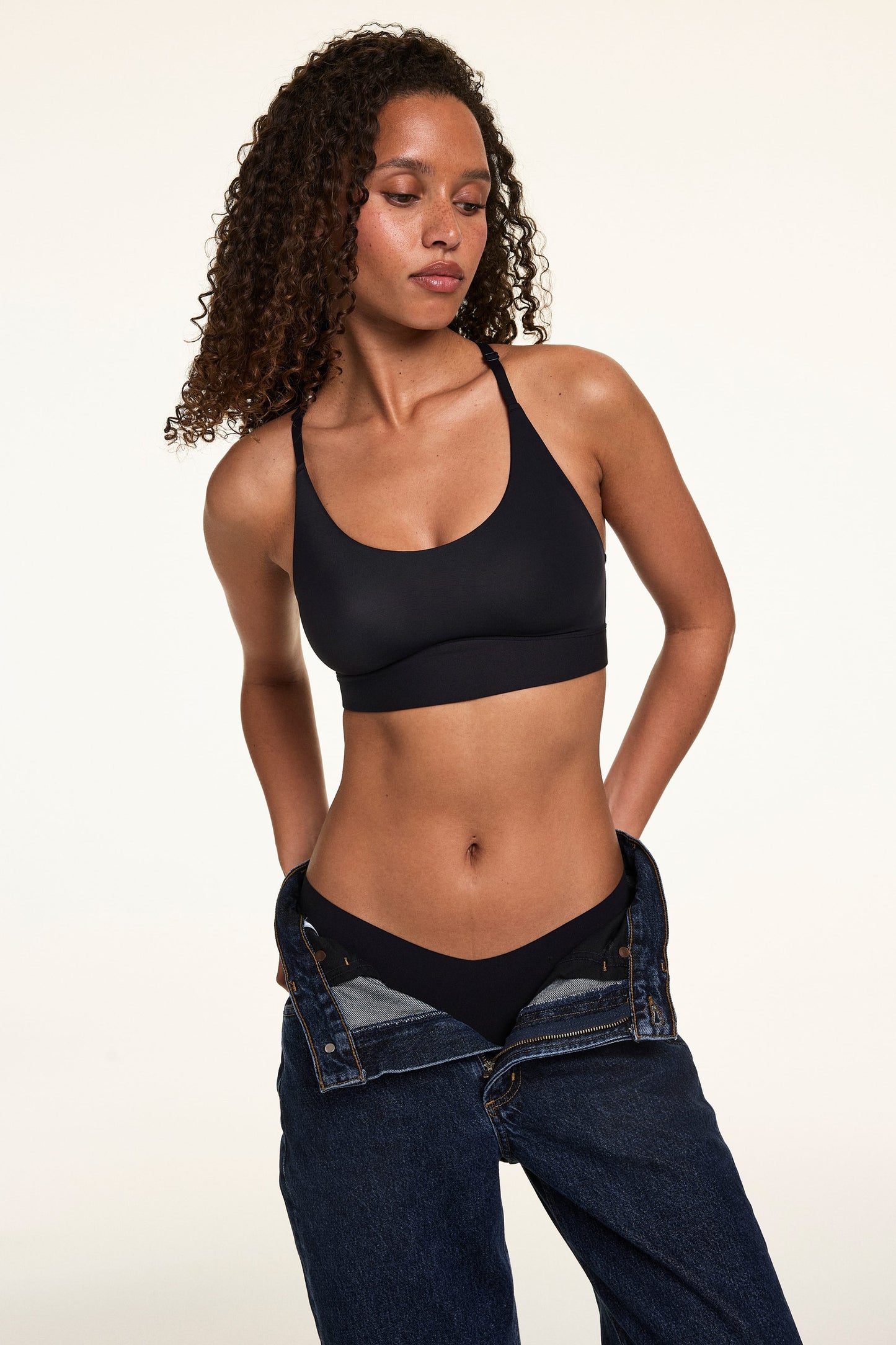 Courtney wears the Bliss Racerback Bralette in Black with matching Black underwear and relaxed-fit jeans, highlighting the racerback silhouette and soft supportive band.