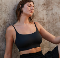 Outdoors against a textured stone wall, a model wears the Bliss Scoop Bralette in Black, styled with black bottoms. The wireless bralette features a scoop neckline, smooth microfiber fabric, and thin adjustable straps designed for soft, everyday comfort.