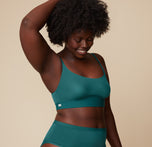 Akesha poses with one hand raised above her head, wearing the Bliss Scoop Bralette in Everglade and highwaist briefs. Her body is angled slightly, emphasizing the bralette’s scoop neckline.