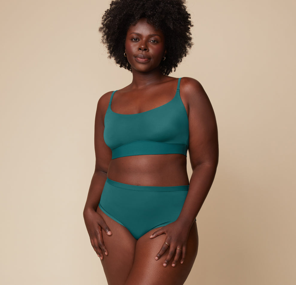 Full-body view of Akesha standing with her hands on her hips, modeling the Bliss Scoop Bralette and matching highwaist briefs in Everglade. The neutral backdrop keeps the focus on her and the fit.