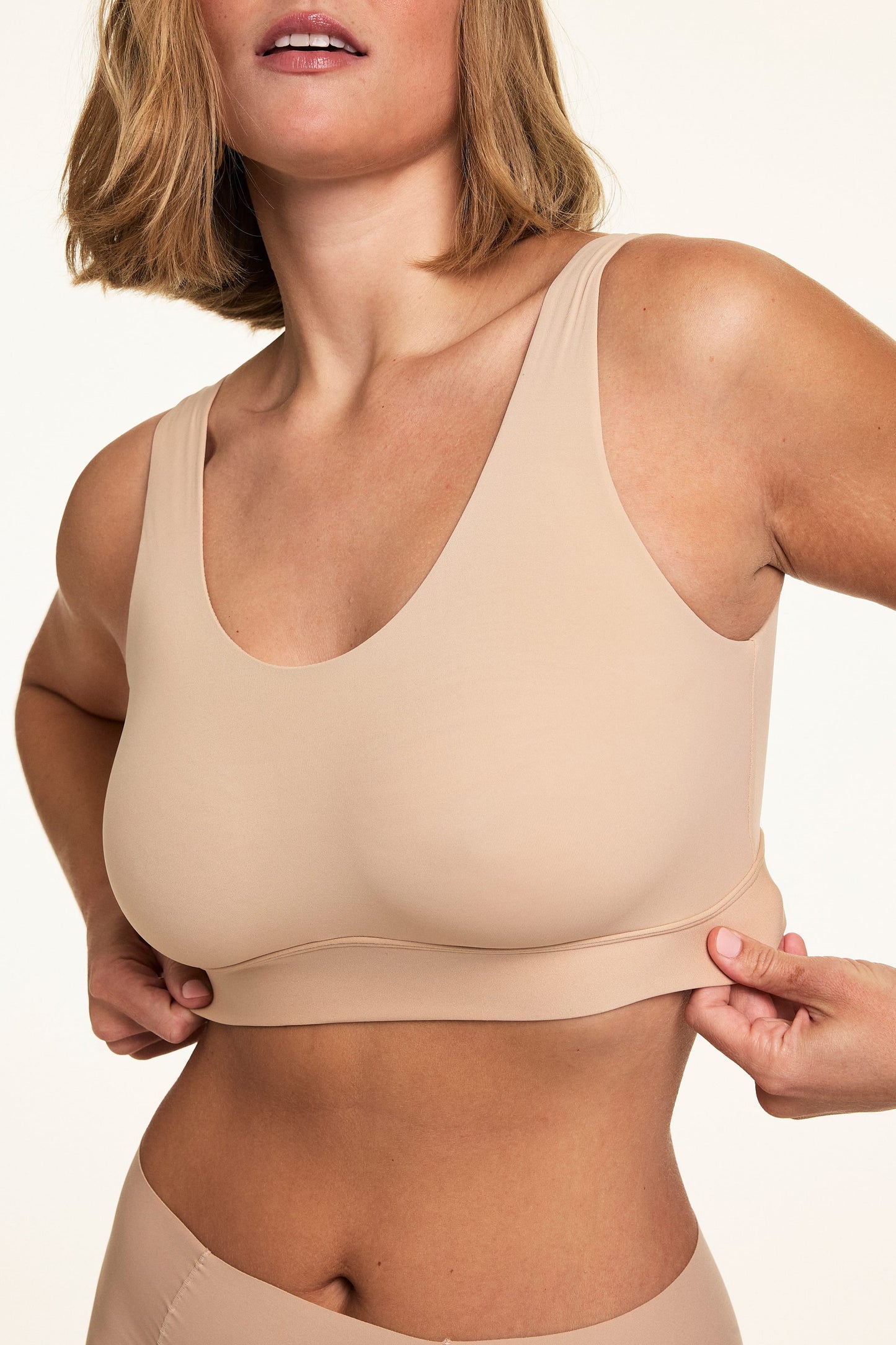 Front view of Sonnie wearing the Bliss Scoop Bralette in Beige, gently pulling the underband to showcase the flexible fit and smooth fabric.