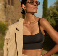 Outdoors in warm sunlight, a model wears the Bliss Square Neck Bralette in Black, showing the square neckline, wireless band, and smooth microfiber fabric. She is styled with tailored pants and a light blazer draped over one shoulder, standing against a stone wall and greenery.
