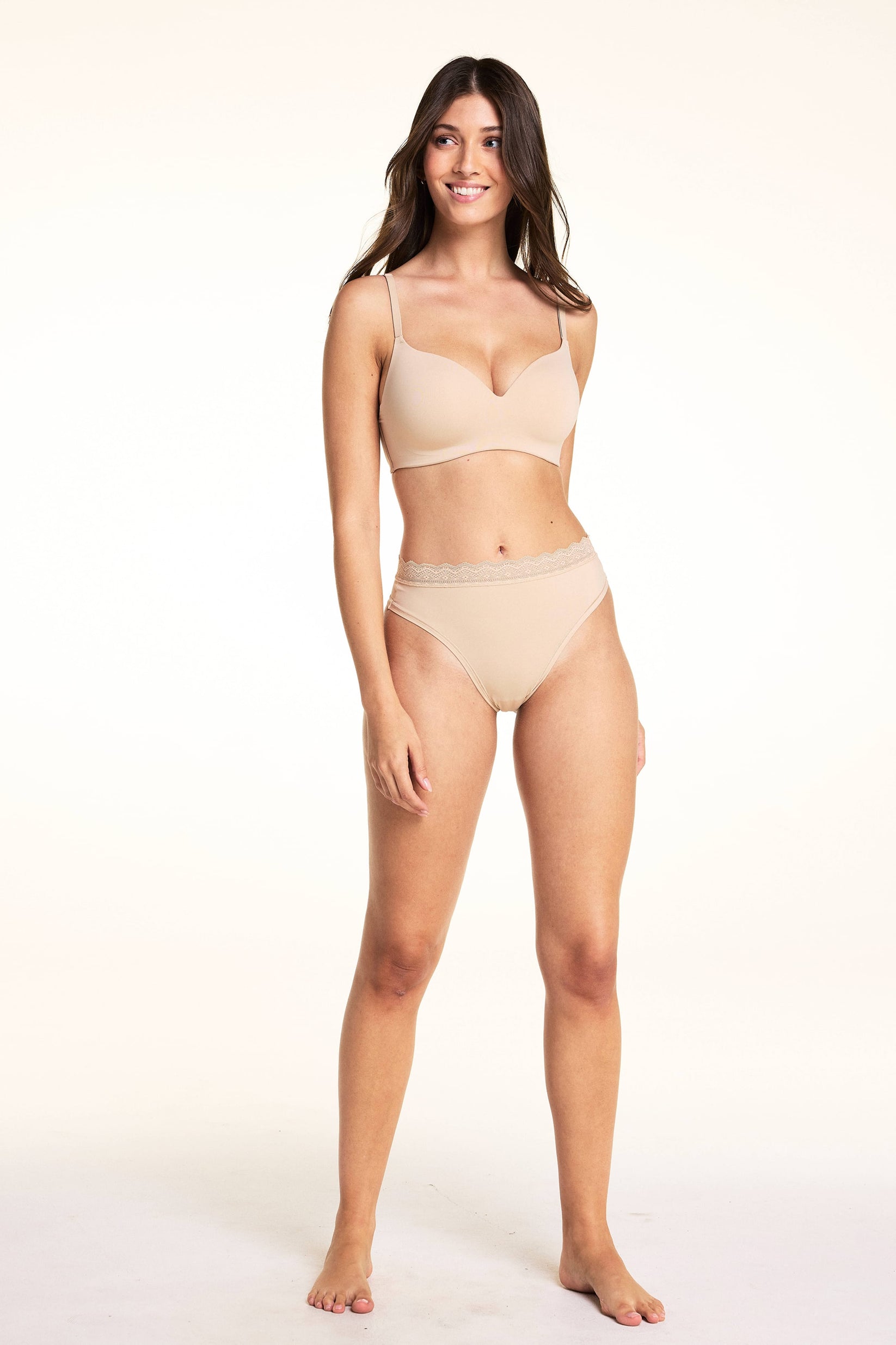 On a light studio backdrop, Laura wears the Bliss Thong in Beige with the Base Wirefree Bra in Beige, smiling in a full-body pose that highlights the smooth finish and lace waistband.