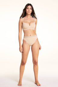 On a light studio backdrop, Laura wears the Bliss Thong in Beige with the Base Wirefree Bra in Beige, standing in a full-body pose to highlight the thong silhouette and lace waistband.