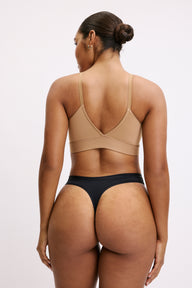 On a white studio backdrop, Amber is shown from the back wearing the Bliss Thong in Black, paired with a wireless bralette in beige. The thong’s minimal back coverage and smooth waistband are visible, designed to sit flat under clothing.