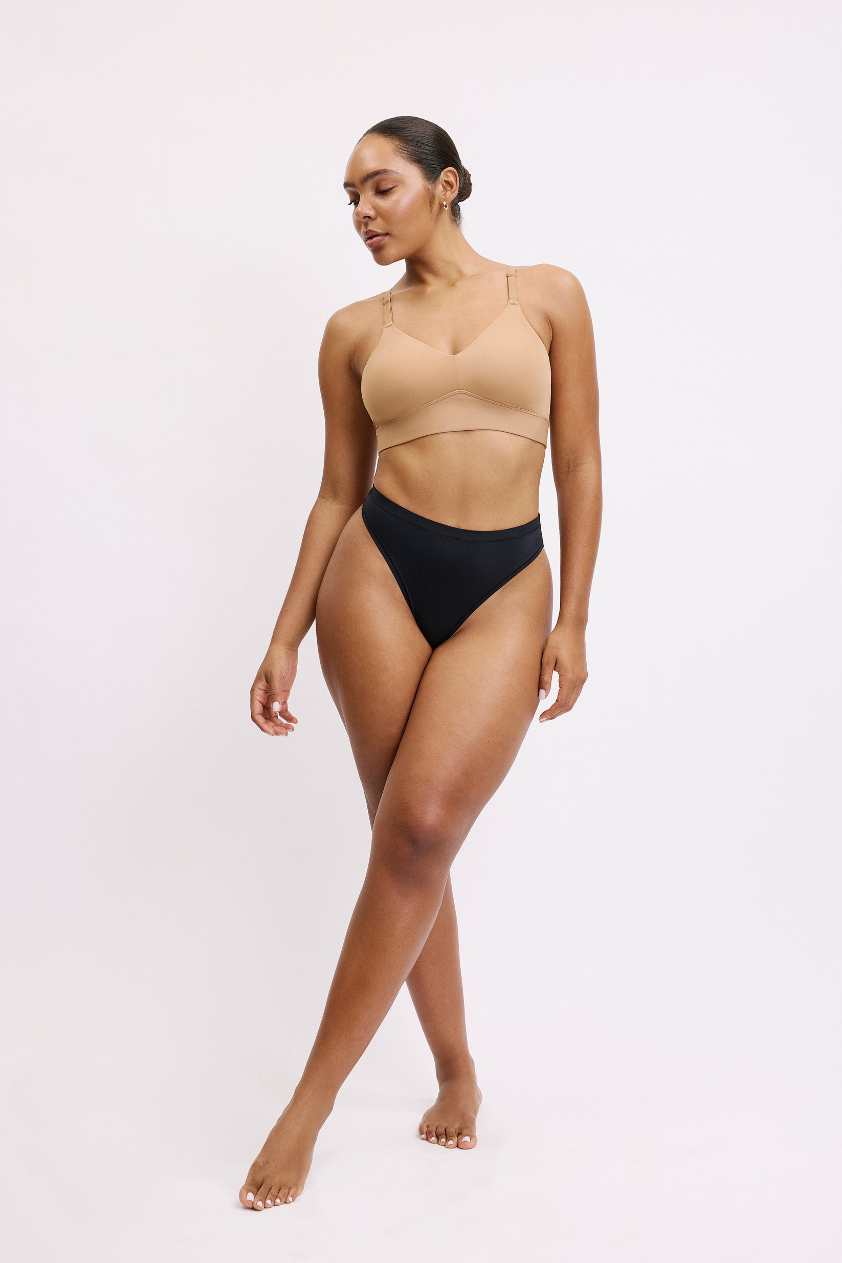 On a white studio backdrop, Amber stands in a three-quarter view wearing the Bliss Thong in Black with a wireless bralette in beige. The thong’s smooth microfiber fabric and clean leg openings are visible for a barely-there feel.