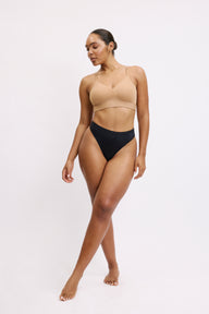 On a white studio backdrop, Amber stands in a three-quarter view wearing the Bliss Thong in Black with a wireless bralette in beige. The thong’s smooth microfiber fabric and clean leg openings are visible for a barely-there feel.