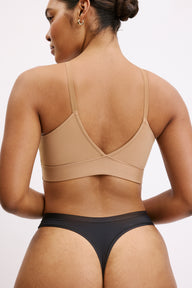 On a white studio backdrop, a close back view shows Amber wearing the Bliss Thong in Black. The single back seam and elastic-free edges are visible, highlighting the no-show design under clothing.