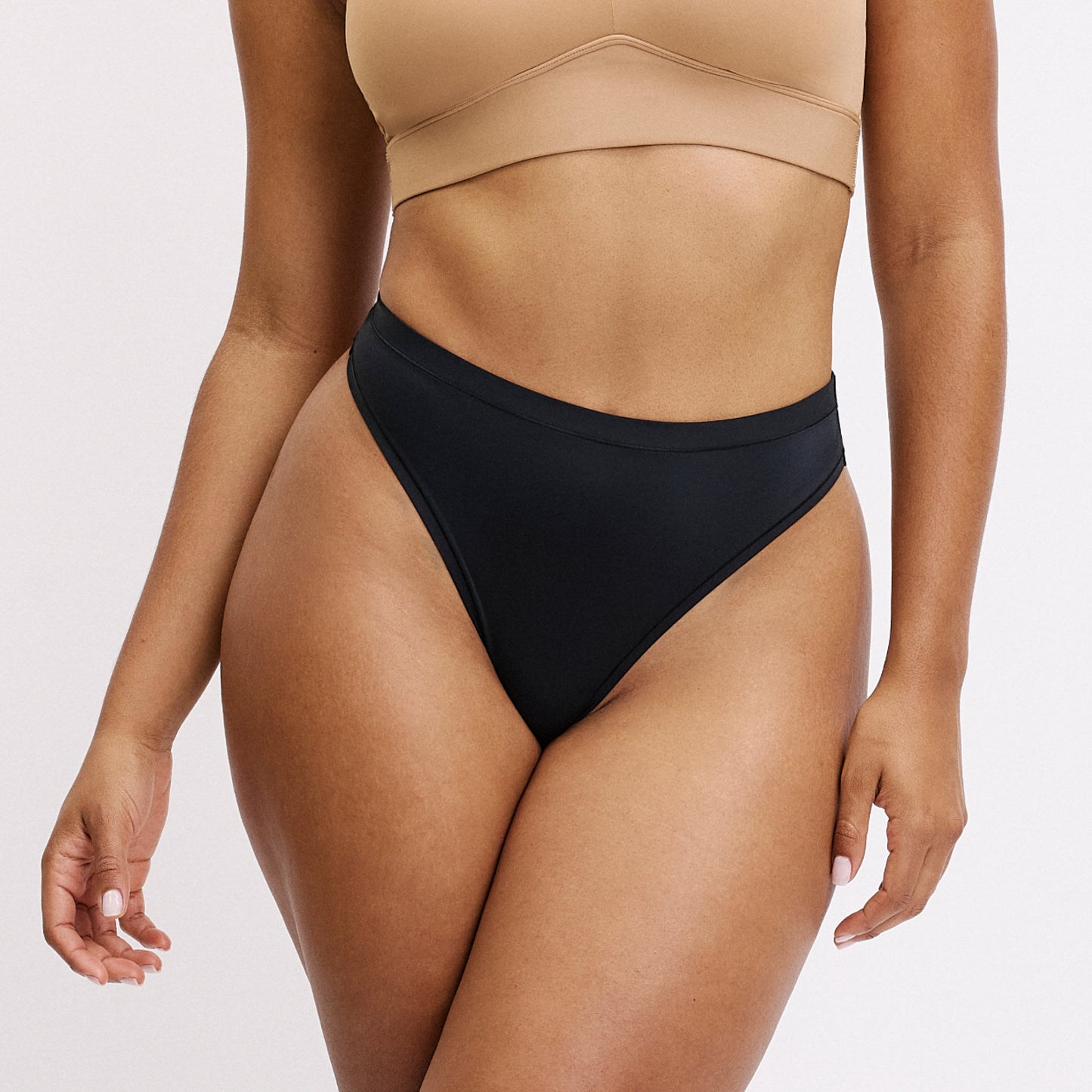 On a white studio backdrop, a close-up shows Amber wearing the Bliss Thong in Black, highlighting the smooth waistband, sleek microfiber fabric, and minimal thong coverage designed to disappear under clothing.