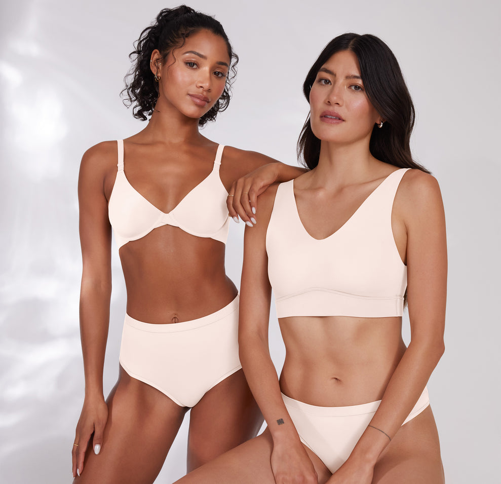 Ally and Anna pose together, both wearing a matching Quartz colored set. Anna wears the Bliss Thong in Quartz and is sitting to showcase the versatile fit. Comfort and inclusivity is emphasized. 