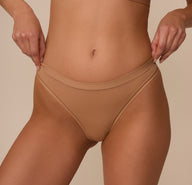 Close-up of Marcela’s hands gently stretching the waistband of the Bliss Thong in Tan, emphasizing the soft stretch and flexible fit.