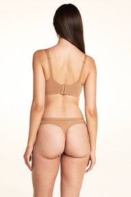 On a light studio backdrop, Laura wears the Bliss Thong in Tan with a matching mesh bra in Tan, standing with her back to the camera to show the thong silhouette and sheer waistband detail.