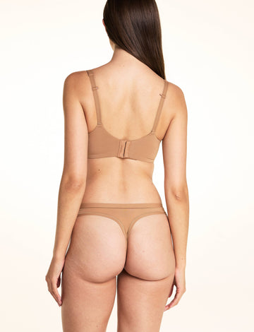 On a light studio backdrop, Laura wears the Bliss Thong in Tan with a matching mesh bra in Tan, standing with her back to the camera to show the thong silhouette and sheer waistband detail.