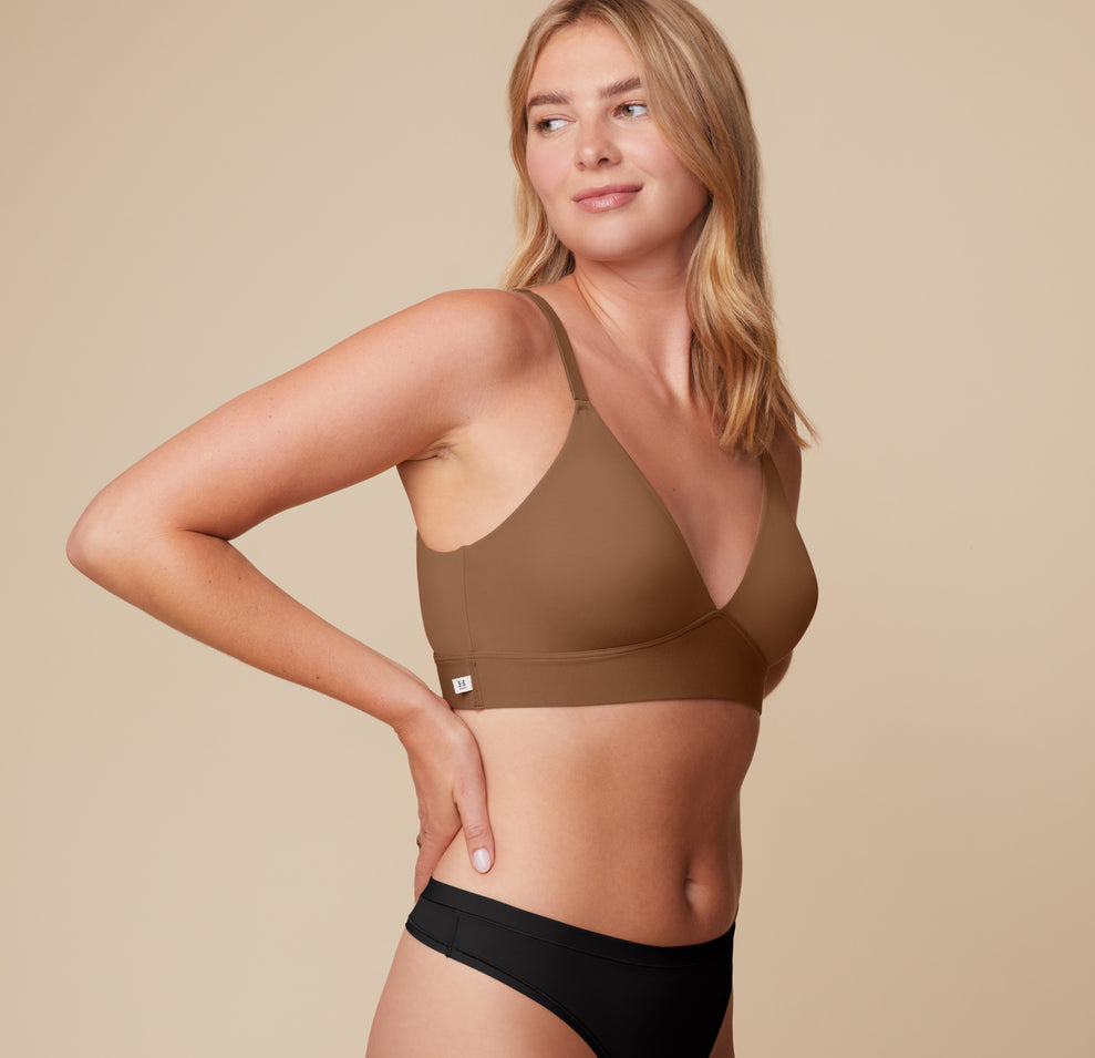 Side profile of Megan turning to look over her shoulder. She wears the Bliss Triangle Bralette in Brown and Bliss Highwaist Thong in Black, emphasizing the bralette’s flattering wrap seam and flexible fit.