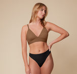 Megan stands with a relaxed pose, one hand on her hip, wearing the Bliss Triangle Bralette in Brown and Bliss Highwaist Thong in Black. The bralette’s deep V-neckline and soft microfiber fabric highlight its comfort-first design.