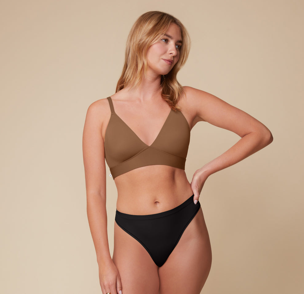 Megan stands with a relaxed pose, one hand on her hip, wearing the Bliss Triangle Bralette in Brown and Bliss Highwaist Thong in Black. The bralette’s deep V-neckline and soft microfiber fabric highlight its comfort-first design.