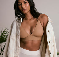 On a soft neutral studio backdrop, a model wears the Bliss Triangle Bralette in Tan, showing the deep V-neckline, smooth microfiber fabric, and wireless band. She layers it with an open light jacket and cream bottoms, standing relaxed with a calm expression.