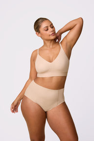 On a white studio backdrop, India wears the Bliss Triangle Bralette in Beige with the matching brief, standing with one arm raised to show the deep V neckline and wireless support.