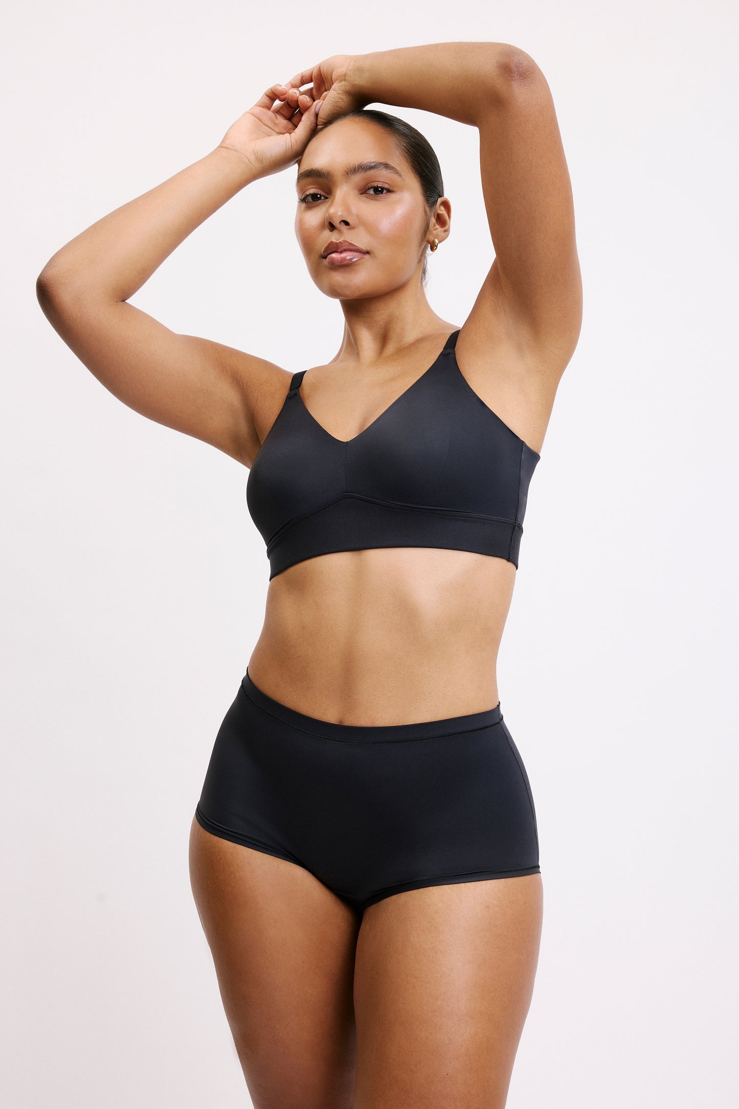 On a white studio backdrop, Amber wears the Bliss Triangle Bralette in Black with arms raised, highlighting the wireless support and clean, minimal silhouette.