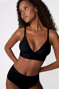 On a white studio backdrop, Courtney wears the Bliss Triangle Bralette in Black, shown from an angled view to highlight the deep V-neckline, smooth microfiber fabric, and front-adjusting straps.