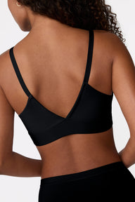 On a white studio backdrop, Courtney is shown from behind wearing the Bliss Triangle Bralette in Black, highlighting the low-back V shape, adjustable straps, and smooth microfiber fabric.