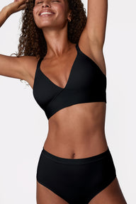 On a white studio backdrop, Courtney wears the Bliss Triangle Bralette in Black with matching bottoms, shown with her arms raised to highlight the deep V-neckline, wireless band, and smooth microfiber fabric. sizing: Courtney wears size Small
