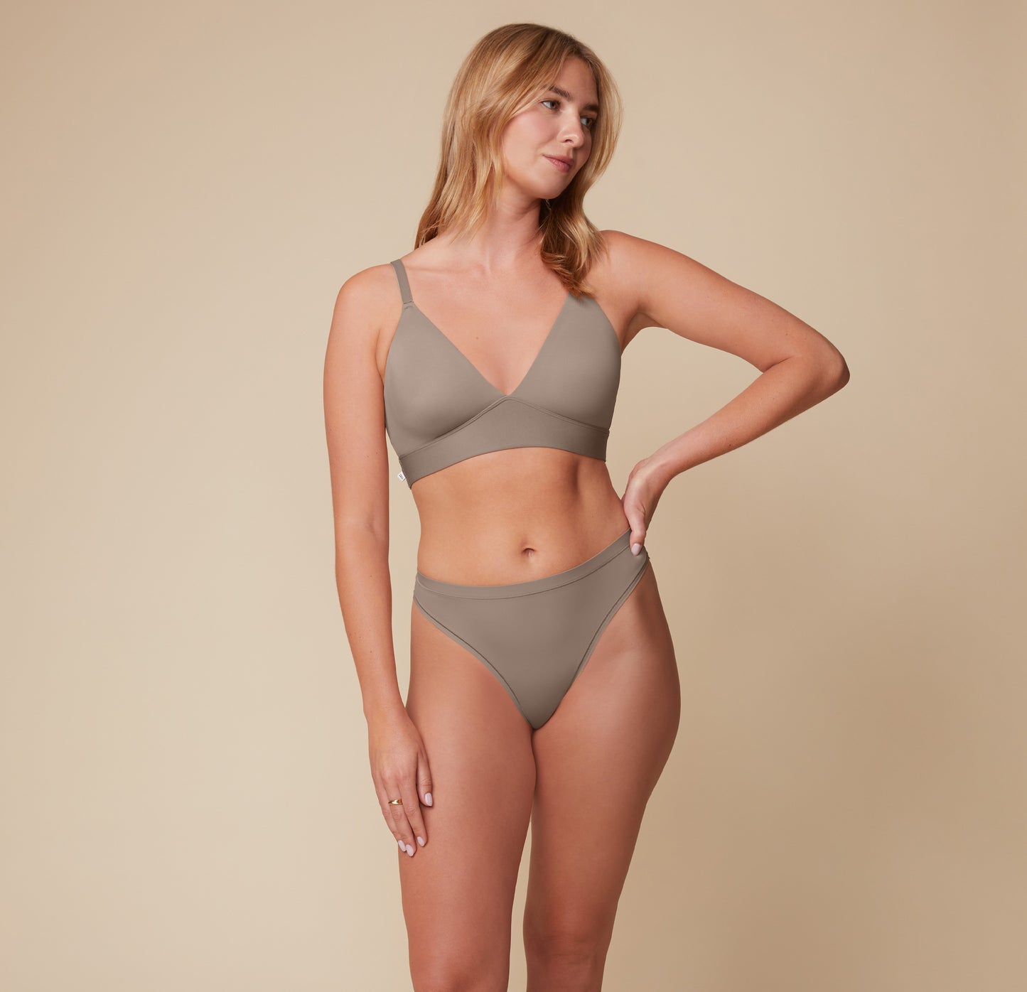 Megan faces forward while looking to the side, wearing the Bliss Triangle Bralette and matching thong in Desert. The smooth fabric and deep V-neckline offer a minimalist, supportive fit.