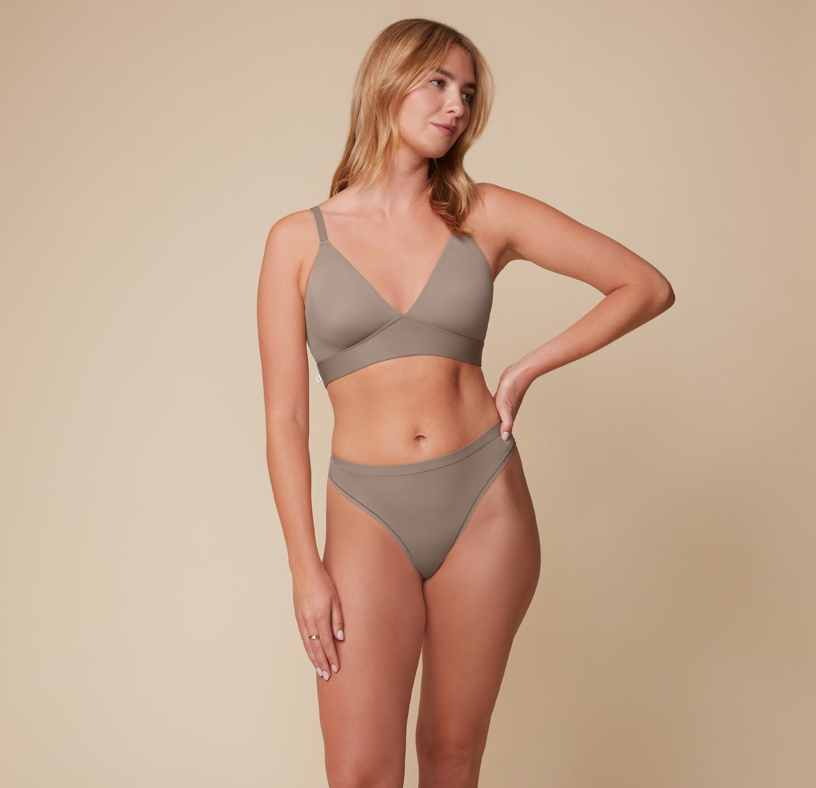 Megan faces forward while looking to the side, wearing the Bliss Triangle Bralette and matching thong in Desert. The smooth fabric and deep V-neckline offer a minimalist, supportive fit.
