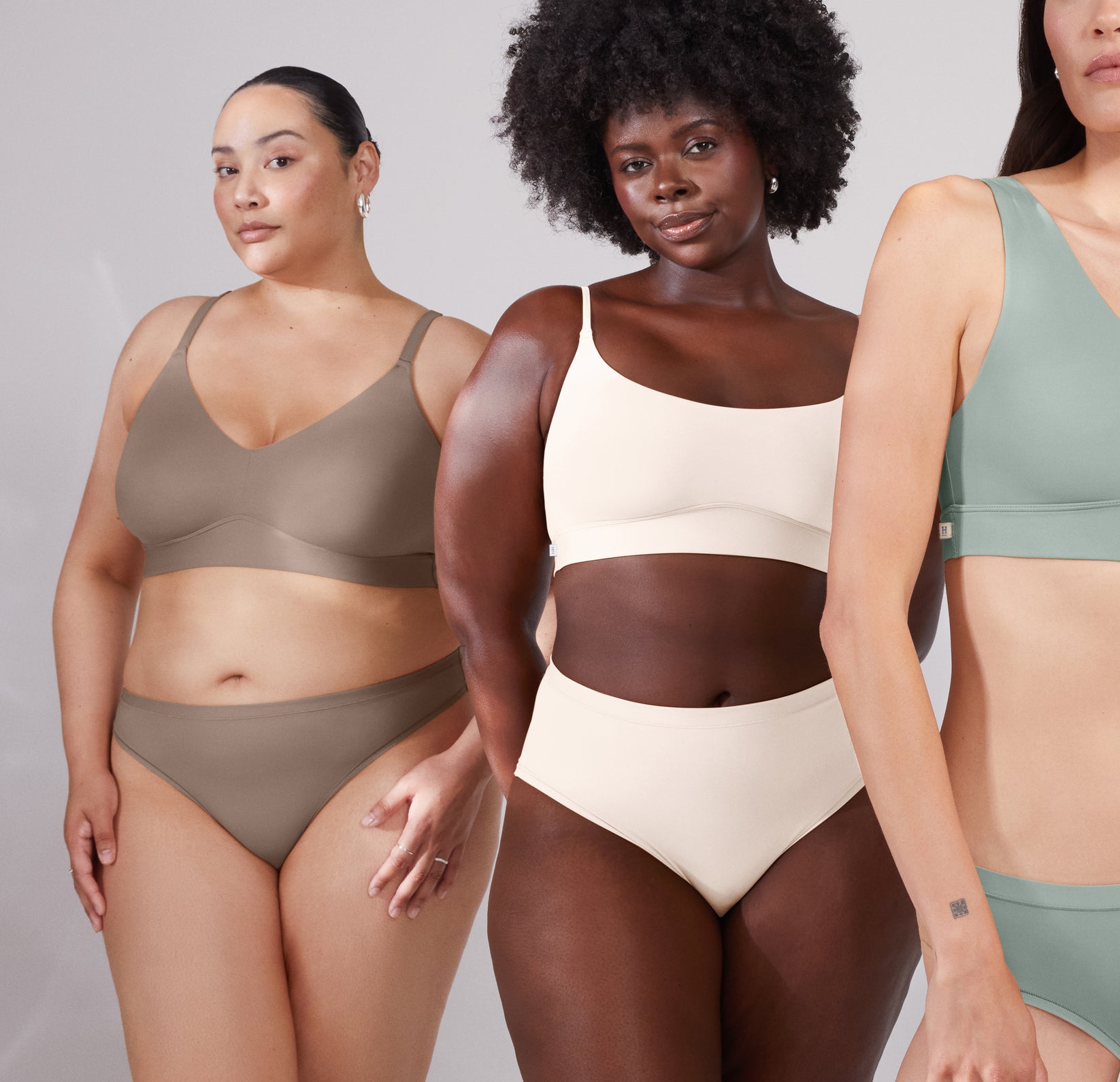 Bianca, Akesha, and Anna pose together in a tiered fashion in the Bliss Triangle Bralette. Bianca wears the color Desert, Akesha wears the color Quartz, and Anna wears the color Seaglass. The image highlights a diverse representation of how the Bliss Triangle Bralette looks on different skin tones and body types. 