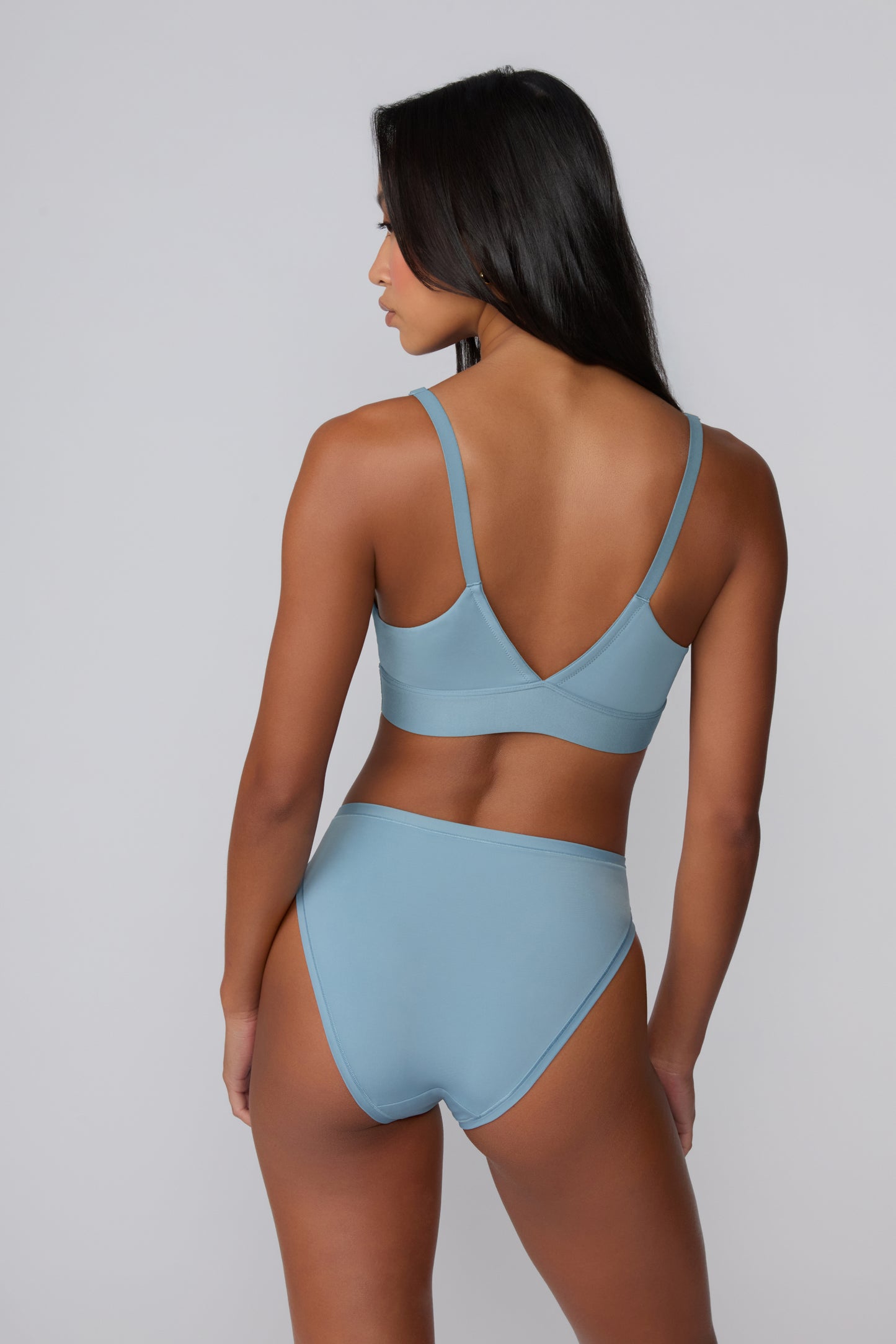 On a light gray studio backdrop, Amber is shown from the back wearing the Bliss Triangle Bralette in Poolside with the matching bikini brief, highlighting the V-shaped back, adjustable straps, and smooth microfiber band.