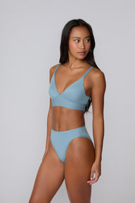 Three-quarter front view on a light gray studio backdrop of Amber wearing the Bliss Triangle Bralette in Poolside with the coordinating bikini brief, showing the deep V neckline, wireless pullover design, and adjustable straps.
