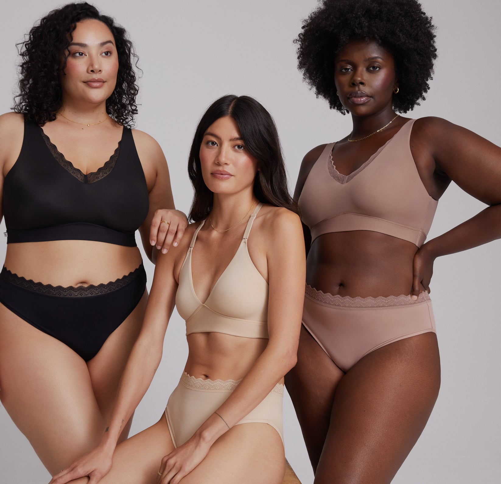 Three models showcasing the Bliss Lace Collection in different shades. From left to right: Bianca wearing the Bliss Lace Bralette and Bliss Lace Highwaist Brief in Black, the second model wearing the Bliss Lace Halterneck Bralette and Bliss Lace Highwaist Brief in Beige, and the third model in the Bliss Lace Bralette and Bliss Lace Highwaist Brief in Mauve. The image highlights the comfortable, supportive fit and lace detailing of the collection, featuring diverse skin tones and body types.