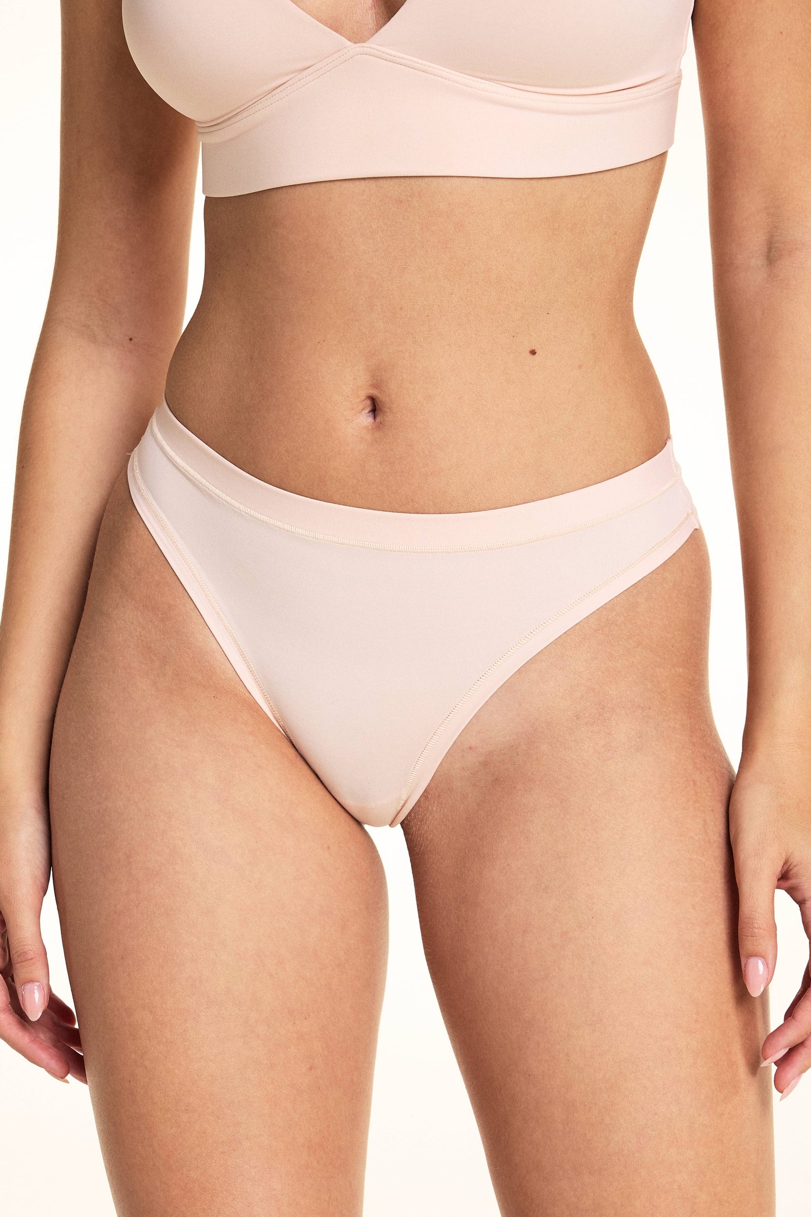 In a studio setting, Laura wears the Bliss Thong in Rose, featuring a smooth microfiber finish, low-rise silhouette, and minimal back coverage with soft elastic trim along the waistband and leg openings. sizing: Rose wears size Small
