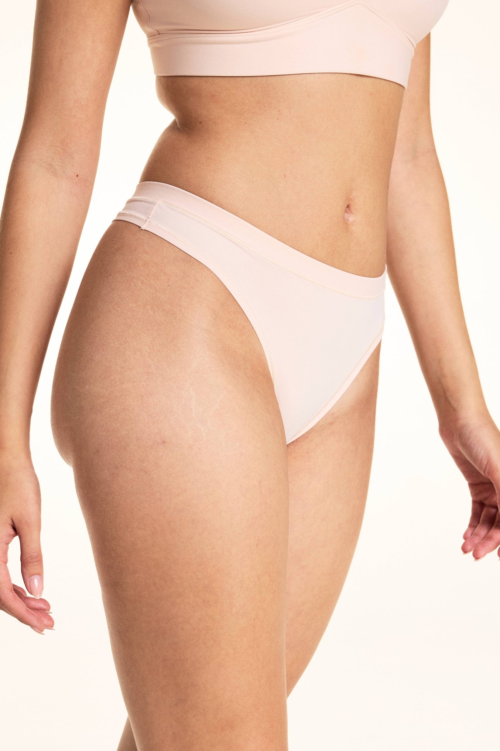 In a studio setting, Laura wears the Bliss Thong in Rose, featuring a smooth microfiber finish, low-rise silhouette, and minimal back coverage with soft elastic trim along the waistband.