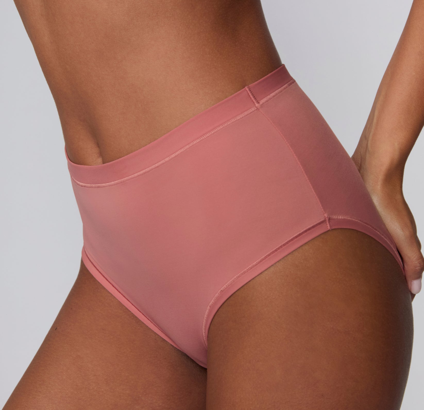 On a light gray studio backdrop, Courtney wears the Bliss Highwaist in Cinnamon, shown from a side angle. The high-waisted underwear features a smooth waistband, soft microfiber fabric, and clean edges designed for comfortable, everyday wear with a no-show feel under clothing.