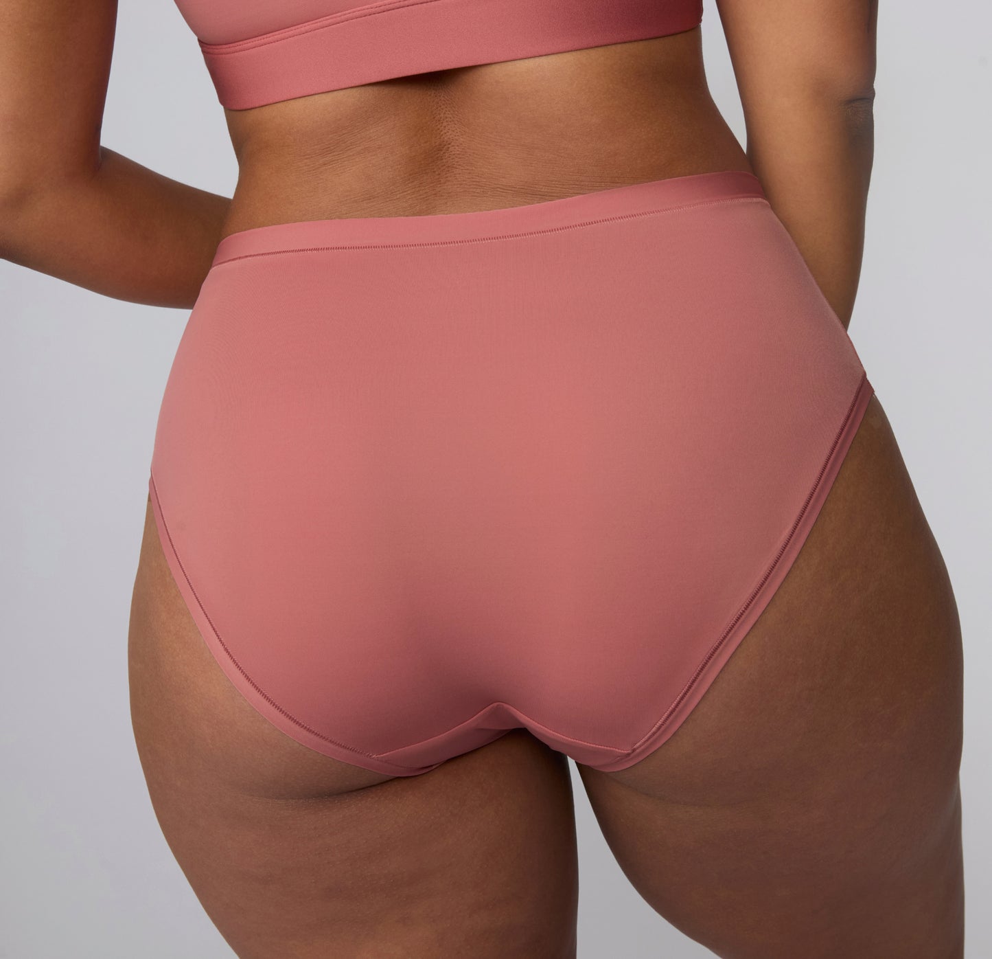 On a light gray studio backdrop, India wears the Bliss Highwaist in Cinnamon, shown from the back. The high-waisted underwear features smooth, full back coverage and soft microfiber fabric designed for comfortable, everyday wear with a no-show feel under clothing.