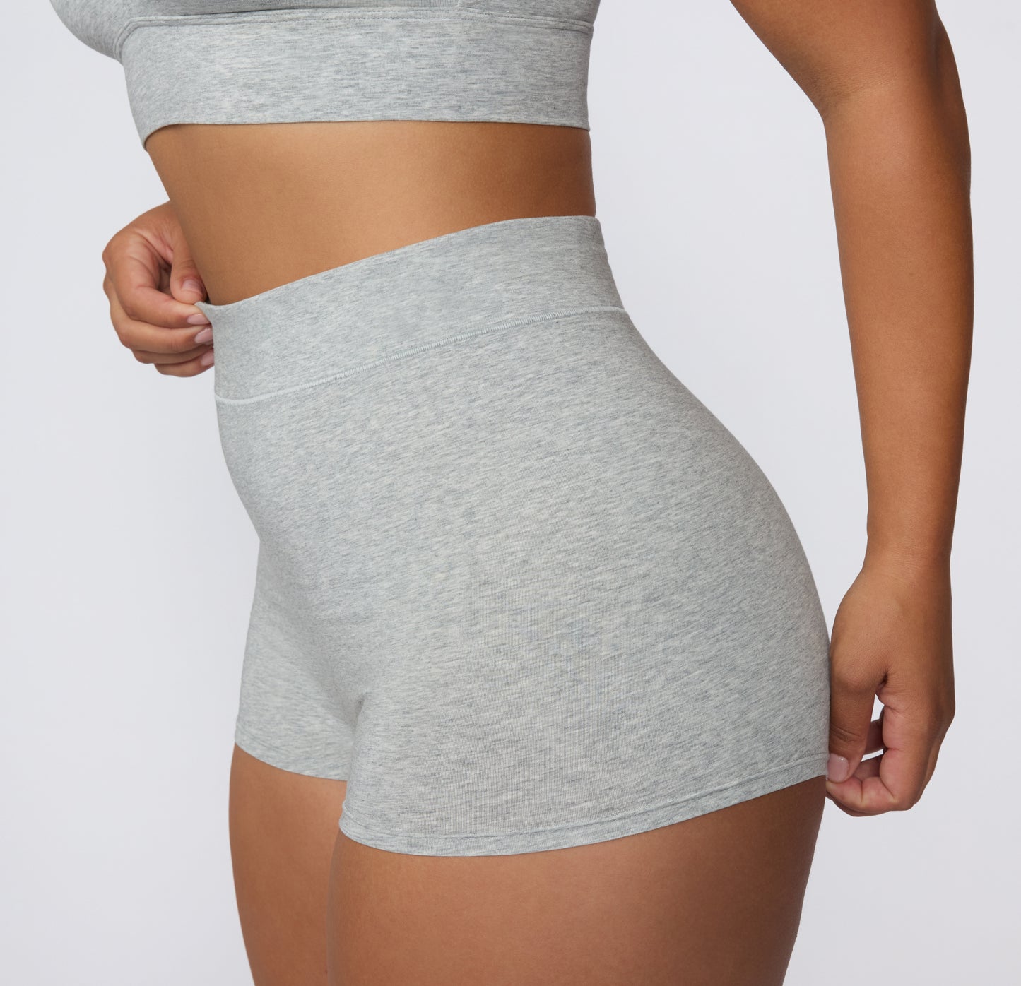 On a white studio backdrop, a model wears the Cloud Cotton Boyshort in Heather Grey, shown from a side angle to highlight the soft, high-rise waistband and smooth cotton fabric.
