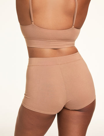 In a softly lit studio, Alana wears the Cloud Cotton Thin Strap Bralette and Cloud Cotton Highwaist Boyshort in Dune, shown from the back. The bralette features thin adjustable straps and a longline band, while the high-waisted boyshort offers full coverage in soft cotton modal fabric.