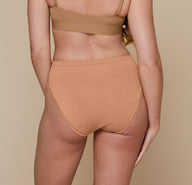 Erin standing with her back to the camera and her arms down by her side.  She’s wearing the Bliss Scoop Bralette in the color Tan and the Cloud Cotton French Cut Brief in the color Dune.