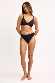 Full-body view on a light studio backdrop of Darion wearing the Cloud Cotton French Cut in Black with a matching black bra. The underwear features a high-rise fit and high-cut leg openings.