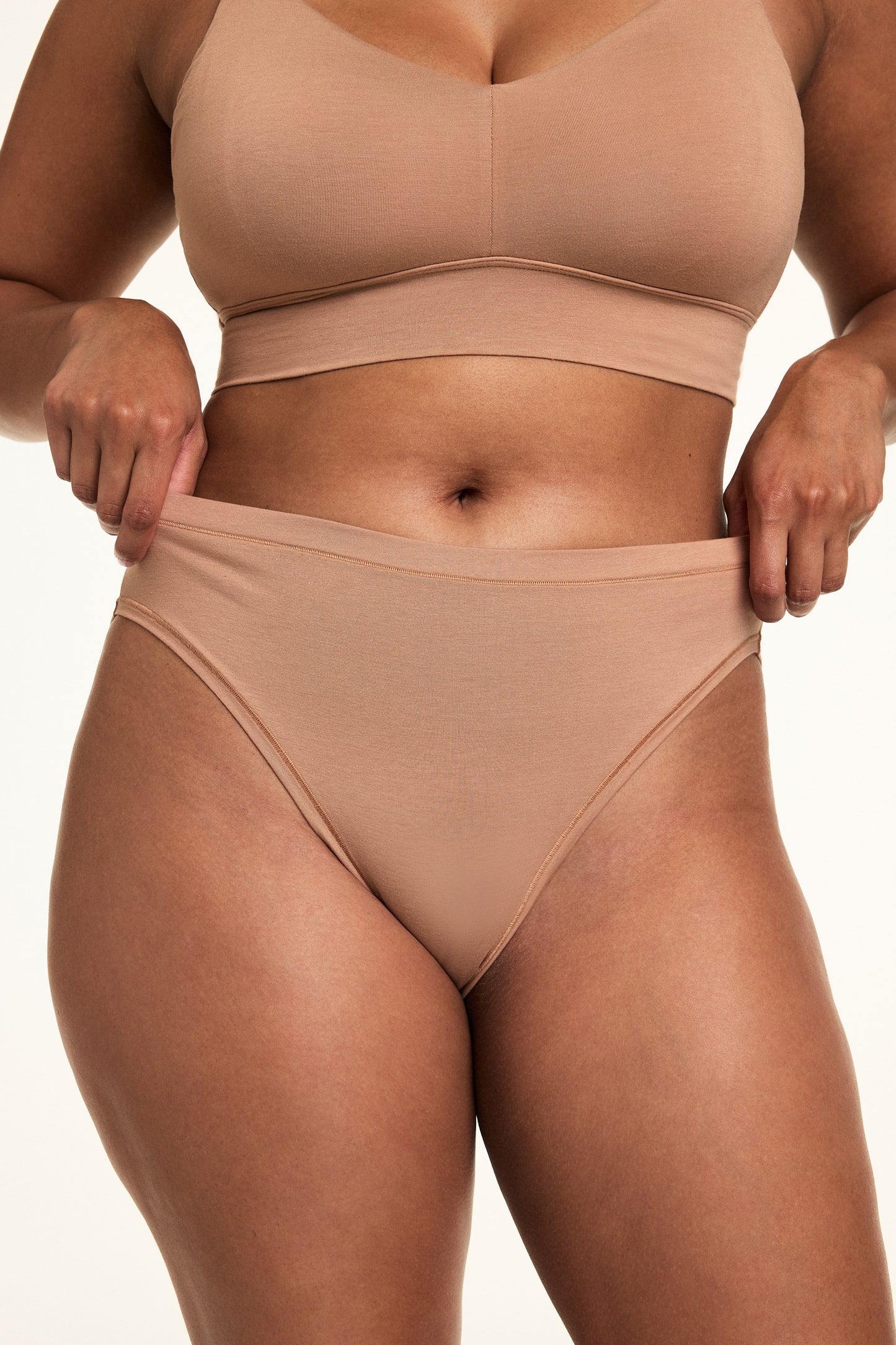 In a softly lit studio, Alana wears the Cloud Cotton Thin Strap Bralette and Cloud Cotton French Cut Brief in Dune. She gently stretches the waistband of the high-rise French cut brief, showing the soft cotton modal fabric and high leg silhouette designed for stay-put comfort.