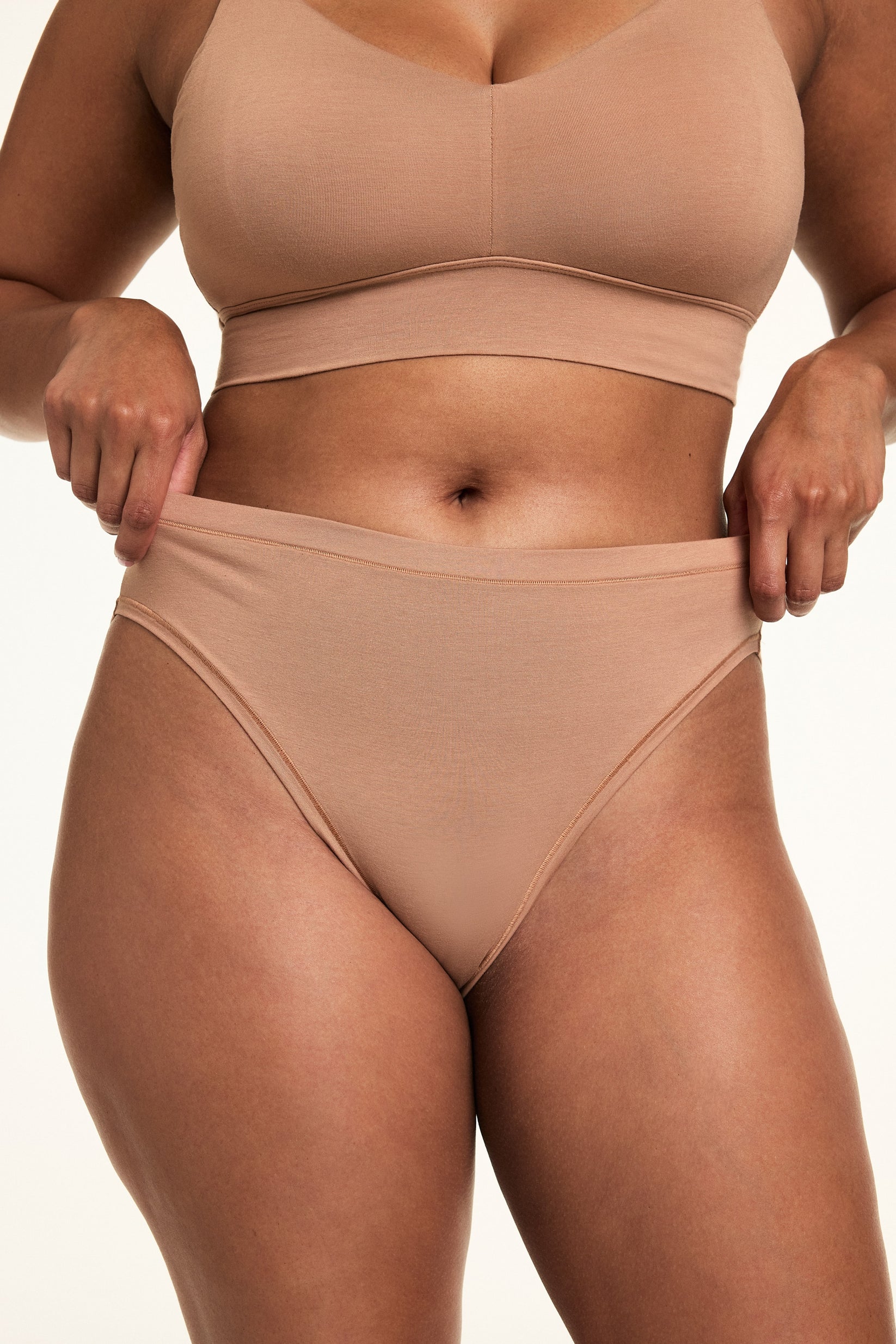 In a softly lit studio, Alana wears the Cloud Cotton Thin Strap Bralette and Cloud Cotton French Cut Brief in Dune. She gently stretches the waistband of the high-rise French cut brief, showing the soft cotton modal fabric and high leg silhouette designed for stay-put comfort.