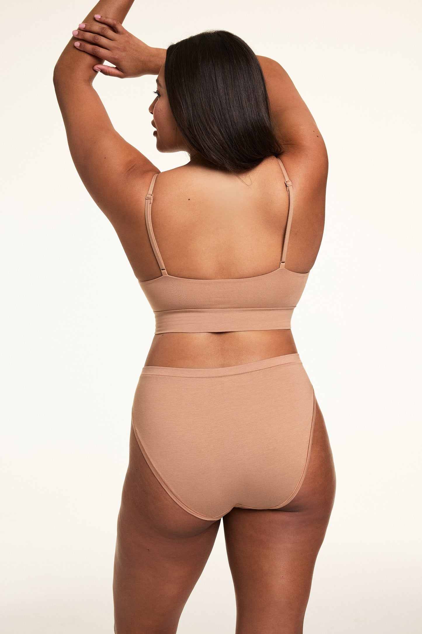 In a softly lit studio, Alana wears the Cloud Cotton Thin Strap Bralette and Cloud Cotton French Cut Brief in Dune, shown from the back. The bralette features thin adjustable straps and a longline silhouette, paired with a high-rise French cut brief in soft cotton modal fabric with moderate back coverage.