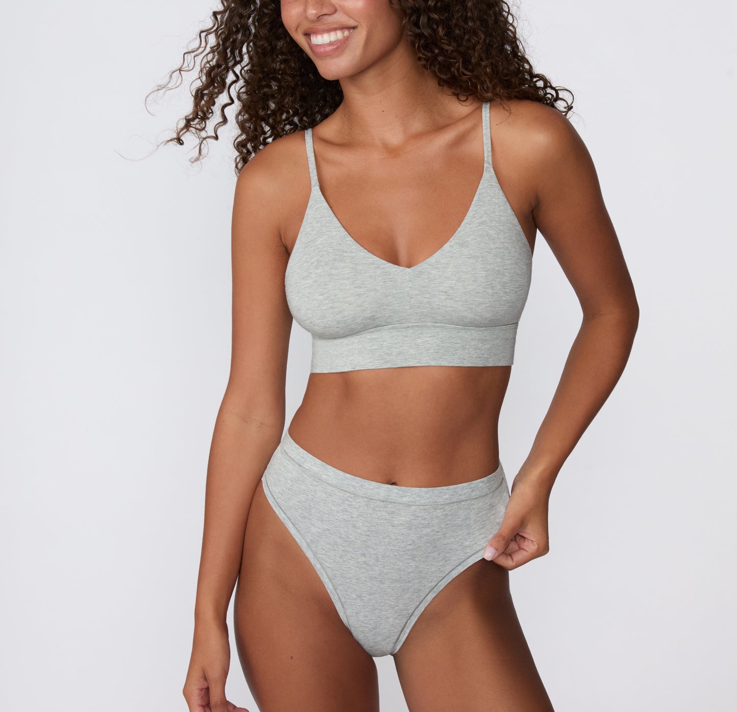 On a white studio backdrop, a model wears the Cloud Cotton Highwaist Thong in Heather Grey with the matching Cloud Cotton Triangle Bralette, showing the V-neckline, thin straps, and high-rise fit.