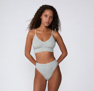 On a white studio backdrop, a model stands wearing the Cloud Cotton Highwaist Thong in Heather Grey with the matching Cloud Cotton Triangle Bralette, showing the high-rise silhouette and lightweight fabric.