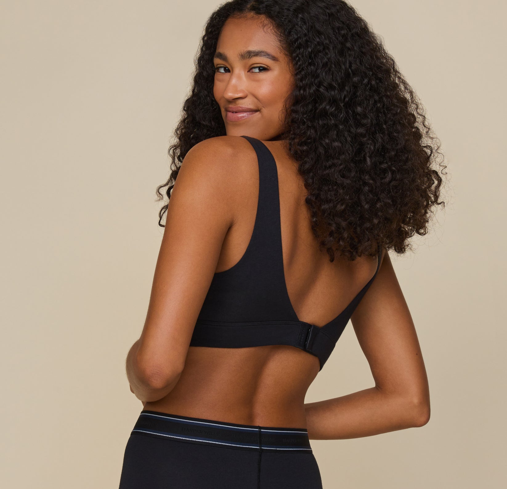 Zoe turns slightly, showing the back of the Cloud Cotton Scoop Bralette in Black. The hook-and-eye closure and deep U-back create a smooth silhouette under clothing.